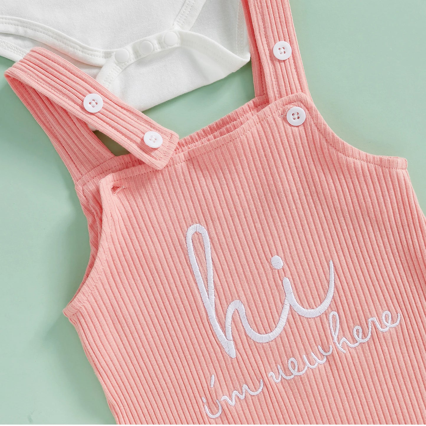 Close-up of baby outfit embroidery reading ‘Hi, I’m new here’ on pink overall